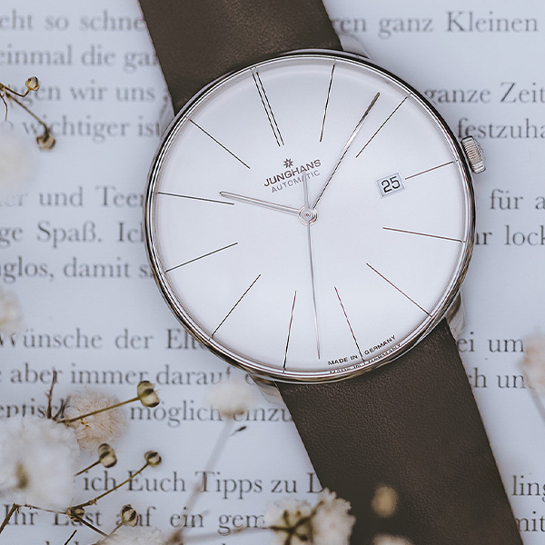 Juwelier-Wagner-Madler-Uhren-Junghans-Kollektion-meister-automatic-ansicht-3 Eine automatische Armbanduhr der Junghans Meister Kollektion mit minimalistischem weißem Zifferblatt und dünnen silbernen Markierungen wird vor dem Hintergrund eines offenen Buches mit deutschem Text präsentiert. Die Uhr mit einem braunen Lederarmband zeigt das Datum auf der 3-Uhr-Position.