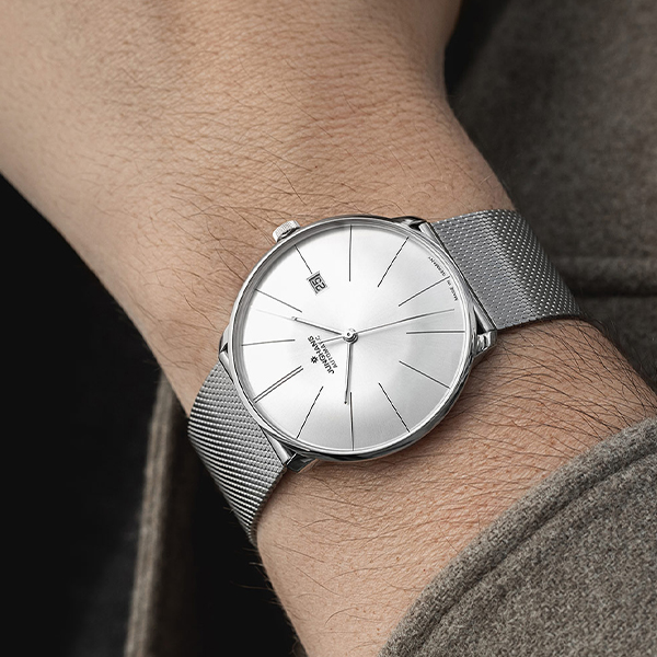 Juwelier-Wagner-Madler-Uhren-Junghans-Kollektion-meister-automatic-ansicht Eine Nahaufnahme eines Handgelenks zeigt eine elegante, silberne Uhr der Junghans Meister Automatik Kollektion mit minimalistischem Design. Die Uhr verfügt über ein kleines Datumsfenster, ein feines Metallarmband und ein einfaches, klares Zifferblatt mit schlanken Stundenmarkierungen und Zeigern. Der Hintergrund enthält einen Teil eines grauen Ärmels.