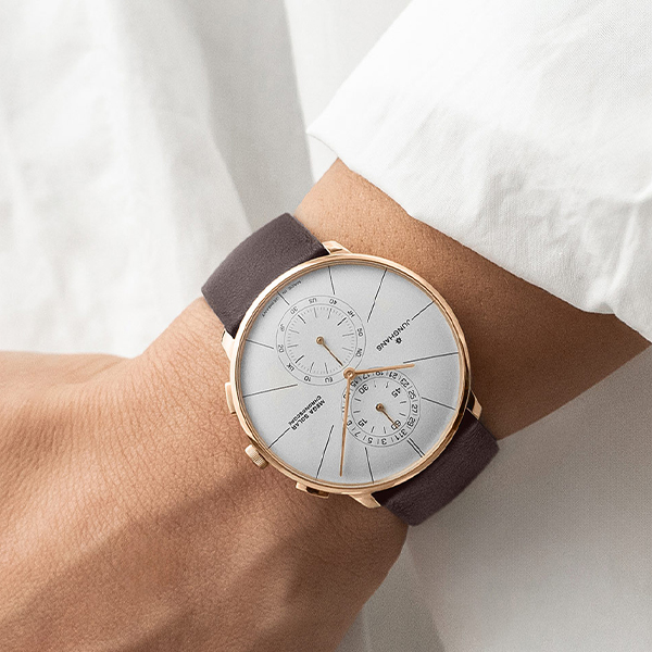 Juwelier-Wagner-Madler-Uhren-Junghans-Kollektion-meister-dunk-und-solar-ansicht-3 Nahaufnahme eines Handgelenks, an dem eine elegante Armbanduhr der Junghans Meister Kollektion mit weißem Zifferblatt und Roségoldgehäuse getragen wird. Die Uhr verfügt über ein schwarzes Lederarmband, zwei Hilfszifferblätter sowie schmale goldene Stundenmarkierungen und Zeiger. Der Träger trägt ein frisches weißes Hemd.
