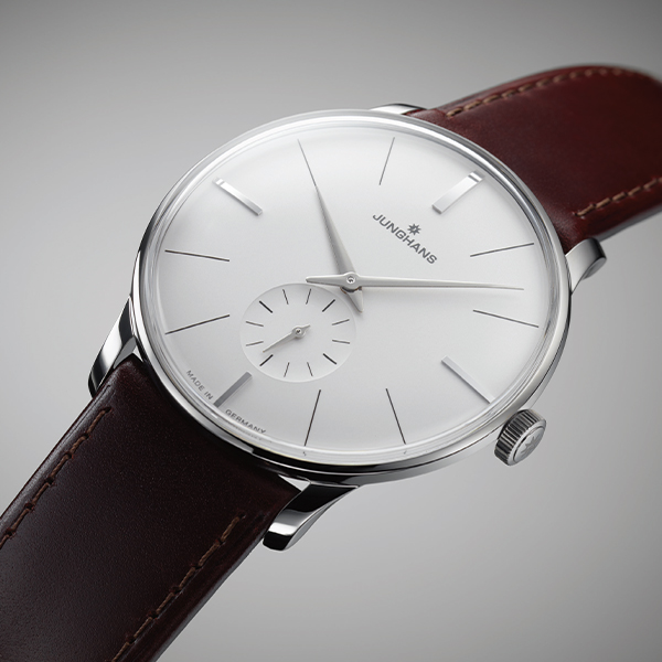 Eine Nahaufnahme einer minimalistischen, silbernen Junghans Meister Handaufzug-Armbanduhr mit weißem Zifferblatt, silbernen Stundenmarkierungen und Zeigern und einem kleinen Sekunden-Hilfszifferblatt auf der 6-Uhr-Position. Die Uhr hat ein braunes Lederarmband und wurde vor einem grauen Hintergrund mit Farbverlauf aufgenommen.