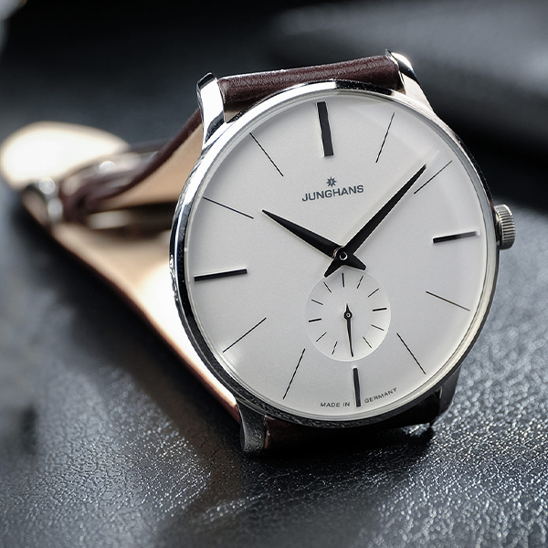 Eine Nahaufnahme einer Junghans Meister Handaufzug-Armbanduhr mit minimalistischem Design. Die Uhr hat ein weißes Zifferblatt, dünne schwarze Stundenmarkierungen und Zeiger, ein kleines Sekundärzifferblatt für die Sekunden auf der 6-Uhr-Position, ein braunes Lederarmband und den Text „MADE IN GERMANY“ auf dem Zifferblatt.