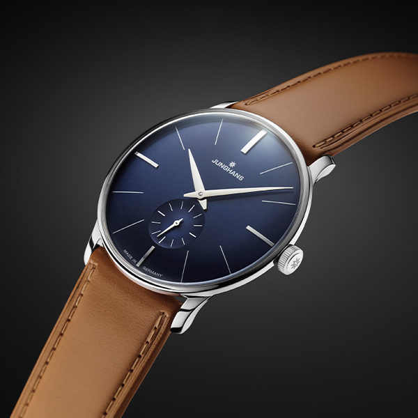 Eine elegante Armbanduhr aus der Junghans Meister Handaufzug Kollektion mit dunkelblauem Zifferblatt, minimalistischem Design, silbernen Markierungen und einem kleinen Hilfszifferblatt bei 6 Uhr. Sie verfügt über ein silbernes Gehäuse und ein braunes Lederarmband, wobei der Markenname „Junghans“ elegant unterhalb der 12-Uhr-Position angebracht ist.