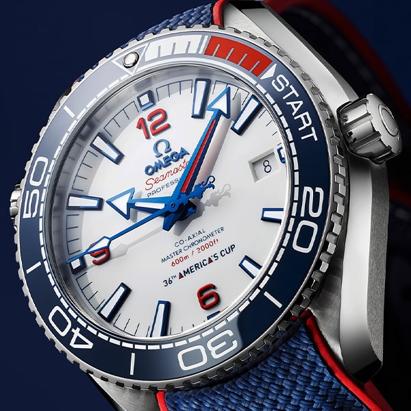 Nahaufnahme einer Omega Seamaster Professional-Uhr aus der Omega Seamaster Kollektion mit weißem Zifferblatt. Blaue Stundenmarkierungen, Zeiger und Lünette ergänzen den roten Minutenzeiger und die „START“-Anzeige auf der Lünette. „36th America’s Cup“ erscheint über 6 Uhr. Das Armband ist blau mit roten Akzenten.