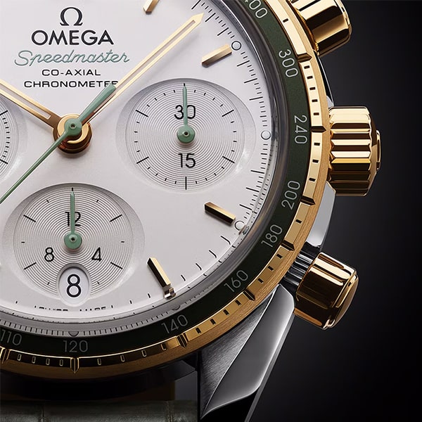Nahaufnahme der Omega Speedmaster Co-Axial Chronometer-Uhr aus der 38er Kollektion. Die Uhr verfügt über ein weißes Zifferblatt mit goldfarbenen Zeigern und Markierungen, drei Hilfszifferblättern sowie goldenen Drückern und einer goldenen Krone. Die Lünette ist dunkel mit einer Tachymeterskala und das Armband scheint aus Leder zu sein.