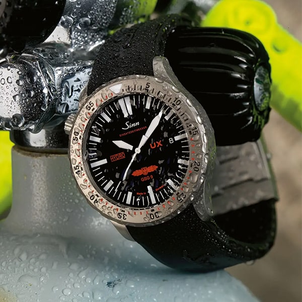 Juwelier-Wagner-Madler-Uhren-Sinn-Ansicht Eine Nahaufnahme einer Sinn-Armbanduhr mit schwarzem Zifferblatt und kräftigen weißen Markierungen sowie einem robusten schwarzen Gummiarmband. Wassertropfen deuten darauf hin, dass sie für nasse Bedingungen geeignet ist, während der Hintergrund, der Tauchausrüstung zeigt, das Tauchthema betont.