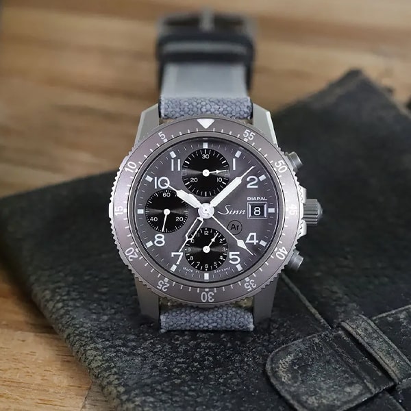 Juwelier-Wagner-Madler-Uhren-Sinn-Kollektionen-103 Ti Diapal Eine Nahaufnahme einer Sinn-Fliegerchronographenuhr aus der Instrumentelle Chronographen Kollektion mit schwarzem Zifferblatt mit mehreren Hilfszifferblättern und robustem grauem Lederarmband. Die Uhr liegt auf einer strukturierten schwarzen Lederoberfläche mit Holzmaserung im Hintergrund.