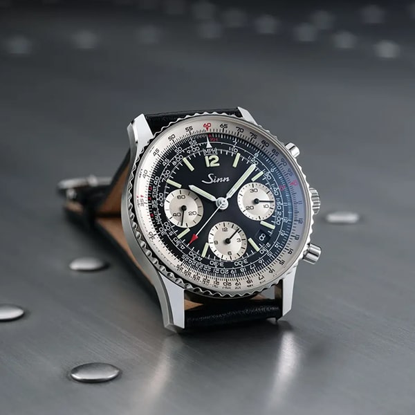 Juwelier-Wagner-Madler-Uhren-Sinn-Kollektionen-Ansicht-2 Eine Nahaufnahme einer Sinn-Armbanduhr zeigt ihr schwarzes Zifferblatt, die silberne Lünette und drei Hilfszifferblätter. Die Uhr verfügt über ein schwarzes Lederarmband und sitzt elegant auf einer mit Nieten verzierten Metalloberfläche. Die Zeit ist 12:15 Uhr.