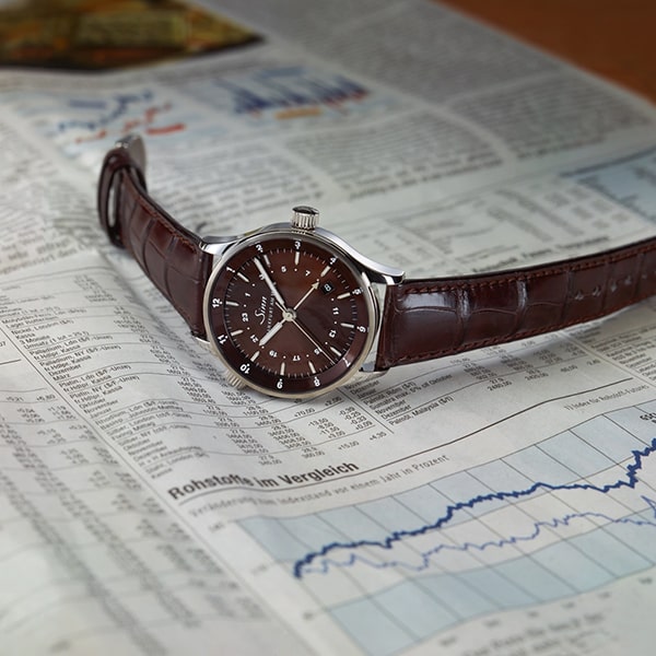 Juwelier-Wagner-Madler-Uhren-Sinn-Kollektionen-Ansicht-6060 Eine stilvolle braune Lederarmbanduhr von Sinn mit silbernem Gehäuse und braunem Zifferblatt liegt auf einer Finanzzeitung mit verschiedenen Diagrammen und Grafiken. Die Uhr ist diagonal über die Seite positioniert, was ihr elegantes Design und ihre detaillierte Verarbeitung unterstreicht.
