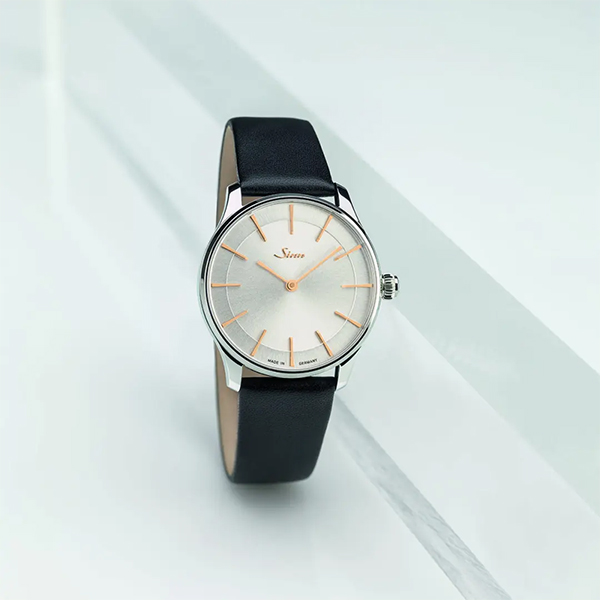 Juwelier-Wagner-Madler-Uhren-Sinn-Kollektionen-Damenuhren-1736stI4N Eine elegante Armbanduhr aus der Sinn Damenuhren Kollektion mit schwarzem Lederarmband und minimalistischem weißen Zifferblatt mit dünnen goldenen Stundenmarkierungen und Zeigern. Das Zifferblatt hat ein kleines Logo und ist in einen glänzenden silbernen Rahmen eingefasst, der auf einem sauberen, hellen Hintergrund mit eckigen Schatten dargestellt wird.