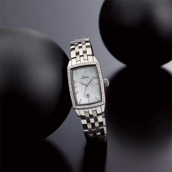 Juwelier-Wagner-Madler-Uhren-Sinn-Kollektionen-Damenuhren-Ansicht-3 Eine polierte silberne Armbanduhr mit rechteckigem Zifferblatt, auf dem der Markenname „Sinn“ aus der Sinn Damenuhren Kollektion zu sehen ist. Die Uhr verfügt über ein Metallarmband mit Gliedern und ist von zwei großen schwarzen Kugeln auf einer reflektierenden schwarzen Oberfläche umgeben.