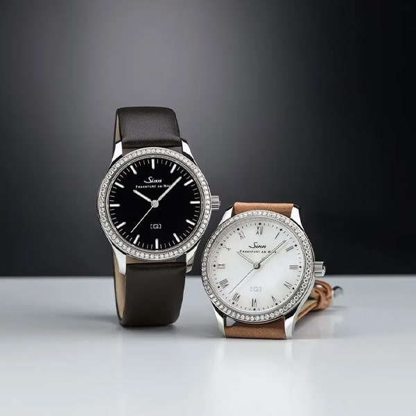 Juwelier-Wagner-Madler-Uhren-Sinn-Kollektionen-Damenuhren-ansicht Vor einem dunklen, unscharfen Hintergrund sind zwei Luxusarmbanduhren aus der Sinn Damenuhren Kollektion zu sehen. Die Uhr links hat ein schwarzes Zifferblatt und ein Lederarmband, während die Uhr rechts ein weißes Zifferblatt und ein hellbraunes Lederarmband hat. Beide haben metallische Lünetten, die mit kleinen Kristallen verziert sind.