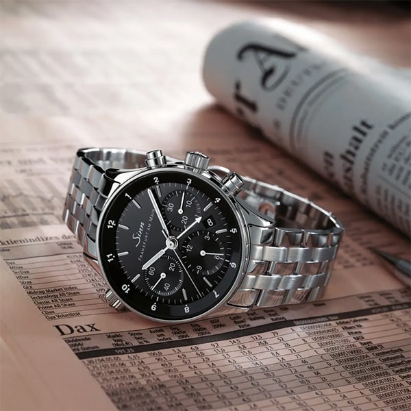 Juwelier-Wagner-Madler-Uhren-Sinn-Kollektionen-Finanzplatz-6000 Eine silberne Armbanduhr aus der Sinn Finanzplatz Kollektion mit schwarzem Zifferblatt und Chronographenfunktion sowie Stahlarmband liegt auf einer Finanzzeitung. Zu sehen ist der Schriftzug „DAX“ und im Hintergrund eine zusammengerollte Zeitung.
