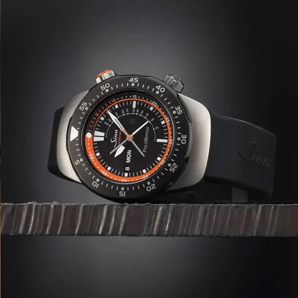 Juwelier-Wagner-Madler-Uhren-Sinn-Kollektionen-Instrumentelle-Chronographen-103 TI diapal Eine Nahaufnahme einer Sinn-Armbanduhr aus der Instrumentelle Chronographen Kollektion mit schwarzem Armband und schlankem, modernem Design. Die Uhr verfügt über ein großes schwarzes Zifferblatt mit weißen und orangefarbenen Akzenten, eine Datums- und Tagesanzeige sowie eine drehbare Lünette mit Minutenmarkierungen. Der dunkle Hintergrund mit Farbverlauf hebt die Uhr hervor.