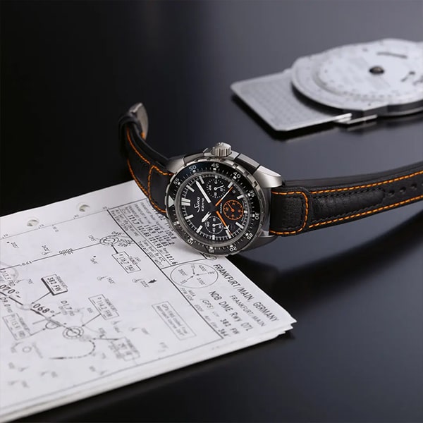 Eine Luxusarmbanduhr aus der Sinn instrumentelle Uhren Kollektion mit schwarzem Lederarmband und orangefarbenen Nähten liegt auf einem technischen Dokument mit Diagrammen und Maßangaben. Im Hintergrund ist ein Rechenschieber auf einer schwarz glänzenden Oberfläche leicht unscharf zu erkennen.