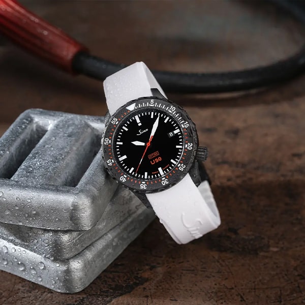 Eine Sinn U50 Armbanduhr aus der Sinn Taucheruhren Kollektion mit schwarzem Zifferblatt und weißem Armband wird auf einem metallischen Objekt präsentiert. Die Uhr verfügt über weiße Stundenmarkierungen, eine Datumsanzeige und eine rote Markierung auf dem Zifferblatt. Sowohl auf der Uhr als auch auf dem metallischen Objekt sind Wassertropfen sichtbar.