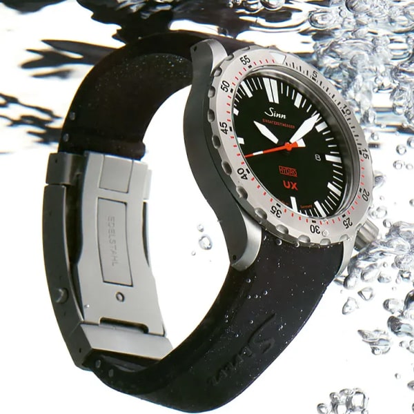 Eine silber-schwarze Taucheruhr aus der Sinn Taucheruhren Kollektion mit Silikonarmband wird in Wasser getaucht. Die Uhr verfügt über ein schwarzes Zifferblatt mit weißen und roten Akzenten und eine drehbare Lünette mit nummerierten Markierungen. Wasserblasen umgeben die Uhr und unterstreichen ihre Unterwasserbeständigkeit.