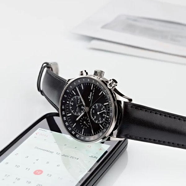 Juwelier-Wagner-Madler-Uhren-Sinn-Kollektionen-finanzplatz Eine stilvolle schwarze Uhr aus der Sinn Finanzplatz Kollektion mit Lederarmband liegt auf einem Smartphone, auf dem eine Kalender-App angezeigt wird. Im Hintergrund sind einige Dokumente teilweise sichtbar. Die Szene ist klar und minimalistisch und unterstreicht die Eleganz der Uhr.