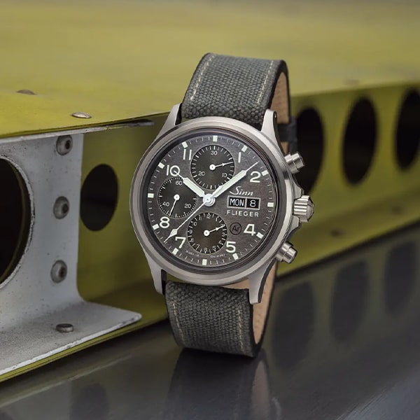 Juwelier-Wagner-Madler-Uhren-Sinn-Kollektionen-instrumentelle-chronographen-ansicht Eine Nahaufnahme einer Sinn Flieger Chronographen-Armbanduhr aus der Sinn Instrumentelle Chronographen Kollektion mit schwarzem Zifferblatt mit weißen Markierungen, drei Hilfszifferblättern und einer Tag-/Datumsanzeige. Die Uhr hat ein Stoffarmband und liegt auf einer industriellen, metallischen Oberfläche mit runden Ausschnitten.