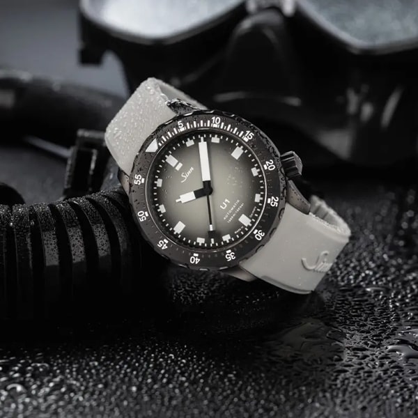 Eine Sinn U1 Taucheruhr aus der Sinn Taucheruhren Kollektion mit weißem Armband und schwarzer Lünette wird vor einem dunklen, nassen Hintergrund präsentiert. Das Zifferblatt verfügt über gut lesbare weiße Stundenmarkierungen und Zeiger, perfekt für den Einsatz unter Wasser. In der Nähe liegt die unverzichtbare Tauchausrüstung, darunter ein schwarzer Schnorchel und eine Maske.