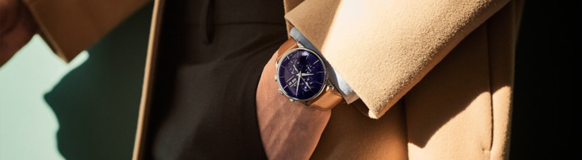 Eine Person in einem beigen Mantel zeigt eine elegante Armbanduhr mit dunkelblauem Zifferblatt und silbernem Gehäuse. Die Uhr hat einfache, elegante Markierungen und Zeiger. Der Hintergrund ist eine grüne Wand, und das Sonnenlicht wirft einen Schatten, der das Zifferblatt hervorhebt.