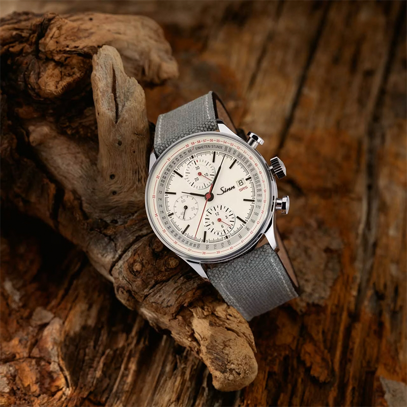 Juwelier-Wagner-Madler-sinn-Kollektionen-Vorschau Eine stilvolle Sinn-Armbanduhr mit grauem Stoffarmband wird vor einem rustikalen Holzhintergrund präsentiert. Die Uhr verfügt über ein silbernes Gehäuse, ein weißes Zifferblatt mit Chronographen-Hilfszifferblättern und eine Datumsanzeige. Der Markenname „Sinn“ ist auf dem Zifferblatt sichtbar.