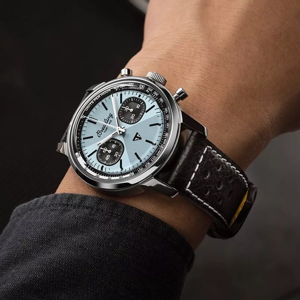 Eine Nahaufnahme eines Handgelenks mit einer Luxusuhr aus der Breitling Top Time Kollektion, mit schwarzem Lederarmband und silbernem Zifferblatt. Die Uhr hat drei Hilfszifferblätter, schwarze Stundenmarkierungen und eine Tachymeterskala um die Lünette. Sie wird mit einem teilweise sichtbaren dunklen Ärmel getragen.