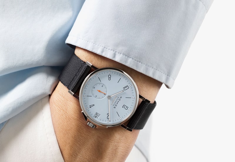 Juwelier-Wagner-Madler-Uhren-Nomos-Glashuette-Ahoi-Kollektion-Ansicht-3 Eine Person in einem hellblauen Hemd präsentiert eine Armbanduhr aus der Nomos Ahoi Kollektion mit einem weißen Zifferblatt mit schwarzen Ziffern und Zeigern. Das elegante Design umfasst ein schwarzes Armband und ein kleines Datumsfenster auf einem schlichten weißen Hintergrund und bietet sowohl Stil als auch Funktionalität.