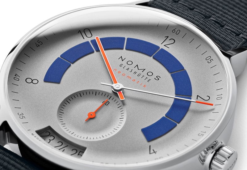 Juwelier-Wagner-Madler-Uhren-Nomos-Glashuette-Autobahn-Kollektion-Ansicht-1 Eine Nahaufnahme einer Uhr von Nomos Glashütte aus der Autobahn Kollektion zeigt ein schlankes, minimalistisches Design mit einem silbernen Zifferblatt und blauen Bögen, die die Stunden markieren. Ein orangefarbener Sekundenzeiger gleitet anmutig über das kleine Hilfszifferblatt bei 6 Uhr und verkörpert moderne Eleganz.