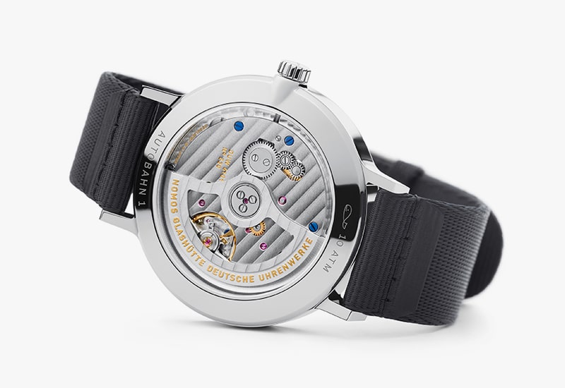 Juwelier-Wagner-Madler-Uhren-Nomos-Glashuette-Autobahn-Kollektion-Ansicht-2B Das Bild zeigt die Rückseite einer Luxusuhr aus der Nomos Autobahn Kollektion mit einem transparenten Gehäuse, das komplizierte mechanische Komponenten offenbart. Die Uhr hat ein schwarzes Armband und die Marke ist dezent um den Gehäuserand eingraviert.