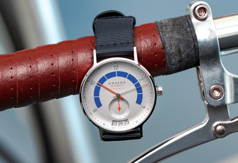 Juwelier-Wagner-Madler-Uhren-Nomos-Glashuette-Autobahn-Kollektion-Ansicht-3 Eine Armbanduhr mit weiß-blauem Zifferblatt aus der Nomos Autobahn Kollektion und schwarzem Armband ist an einem mit rotem Leder umwickelten Fahrradlenker befestigt. Die Uhr zeigt unten das Datum an und trägt stolz den Markennamen „Nomos“ auf dem Zifferblatt.