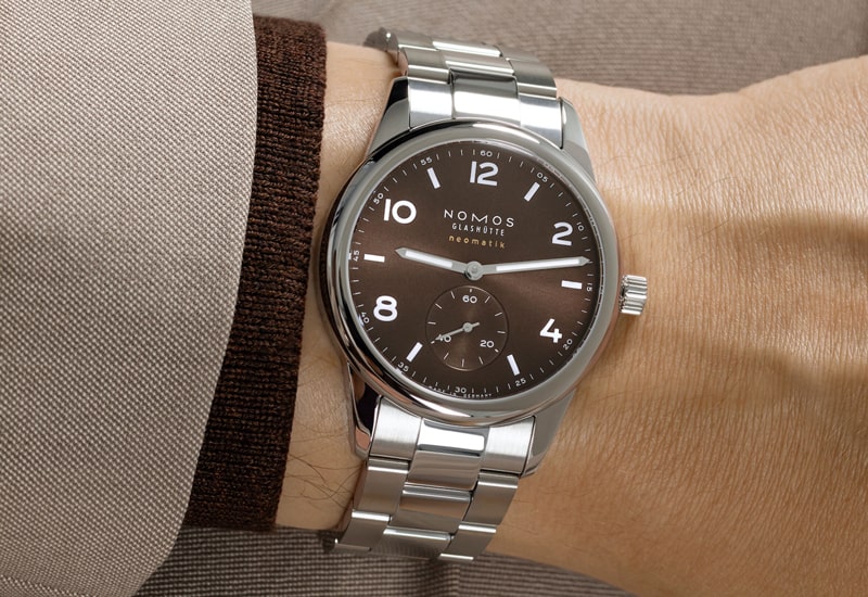 Juwelier-Wagner-Madler-Uhren-Nomos-Glashuette-Club-Kollektion-Ansicht-1 Eine Armbanduhr aus der Nomos Club Kollektion verfügt über ein braunes Zifferblatt mit kleiner Sekunde, weißen Ziffern und Zeigern. Das metallische Armband ergänzt das Design, während „NOMOS“ deutlich sichtbar unterhalb der 12-Uhr-Position an Ihrem Handgelenk angezeigt wird.