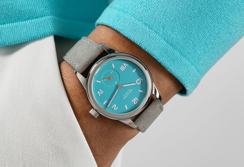 Juwelier-Wagner-Madler-Uhren-Nomos-Glashuette-Club-Kollektion-Ansicht-2 Eine Person trägt einen türkisfarbenen Pullover und eine hellgraue Hose und trägt eine Armbanduhr aus der Nomos Club Kollektion am Handgelenk. Die Uhr hat ein türkisfarbenes Zifferblatt mit silbernen Zeigern und ein hellgraues Armband.