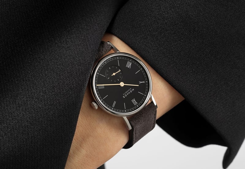 Juwelier-Wagner-Madler-Uhren-Nomos-Glashuette-Ludwig-Kollektion-Ansicht-1 Eine Person trägt eine elegante schwarze Uhr aus der Nomos Ludwig Kollektion mit rundem silbernem Gehäuse und braunem Lederarmband. Das Zifferblatt zeigt römische Ziffern, schmale Zeiger und ein zusätzliches kleines Zifferblatt für die Sekunden. Die Person trägt ein schwarzes Outfit.