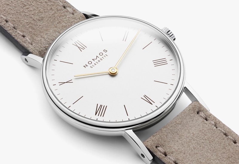Juwelier-Wagner-Madler-Uhren-Nomos-Glashuette-Ludwig-Kollektion-Ansicht-2 Eine Nahaufnahme einer Uhr der Nomos Ludwig Kollektion mit weißem Zifferblatt und römischen Ziffern. Die Uhr verfügt über schlanke goldene Zeiger und ein beiges Wildlederarmband und verkörpert zeitlose Eleganz.