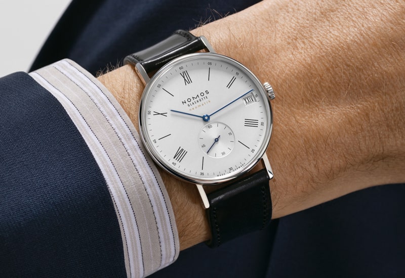 Juwelier-Wagner-Madler-Uhren-Nomos-Glashuette-Ludwig-Kollektion-Ansicht-3 Nahaufnahme einer Person, die eine Nomos Glashütte-Uhr aus der Nomos Ludwig Kollektion trägt. Die Uhr hat ein weißes Zifferblatt, römische Ziffern und ein Datumsfenster. Sie verfügt über ein kleines Sekunden-Hilfszifferblatt und ein schwarzes Lederarmband. Die Person ist elegant gekleidet und trägt einen Anzug mit blauem Hemd.