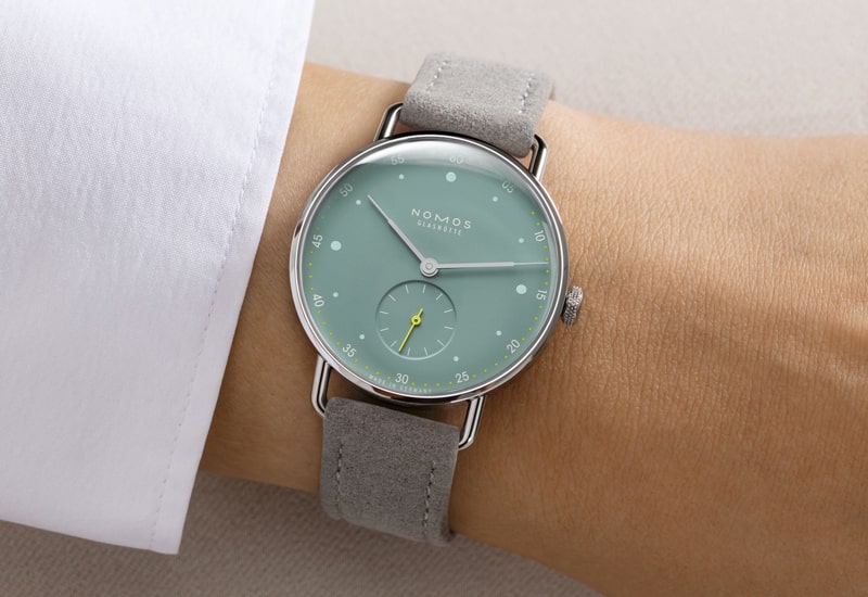 Eine Armbanduhr aus der Nomos Metro Kollektion hat ein hellgrünes Zifferblatt, minimalistisches Design und ein silbernes Gehäuse. Sie schmückt das Handgelenk mit Eleganz, verfügt über ein kleines Hilfszifferblatt und ein hellgraues Wildlederarmband, das unter einem frischen weißen Hemdärmel hervorschaut.