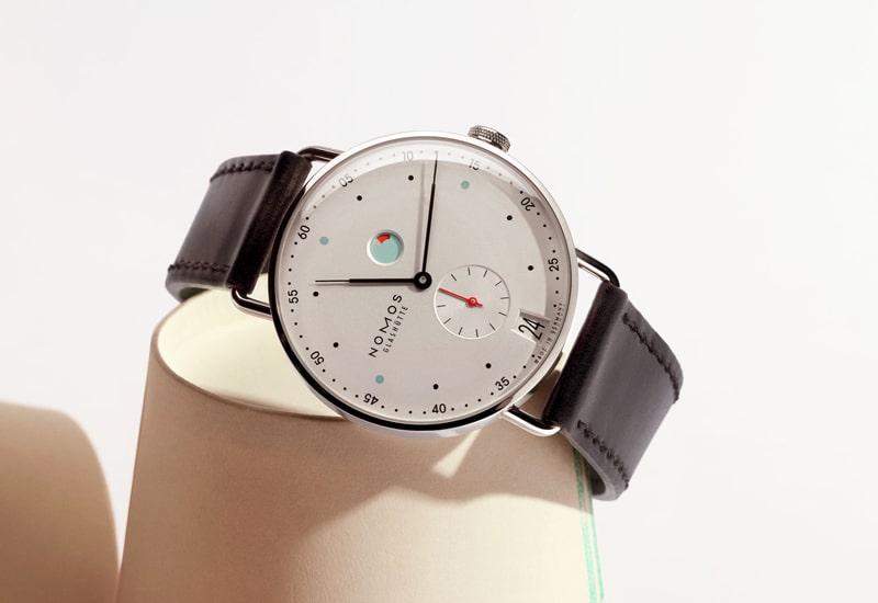 Eine Armbanduhr aus der Nomos Metro Kollektion, die ein minimalistisches Design mit weißem Zifferblatt, eleganten schwarzen Stundenmarkierungen, einem Datumsfenster und Hilfszifferblättern hervorhebt. Sie verfügt über ein silbernes Gehäuse und ein schwarzes Lederarmband, das sich elegant vor einem hellen Hintergrund abhebt.