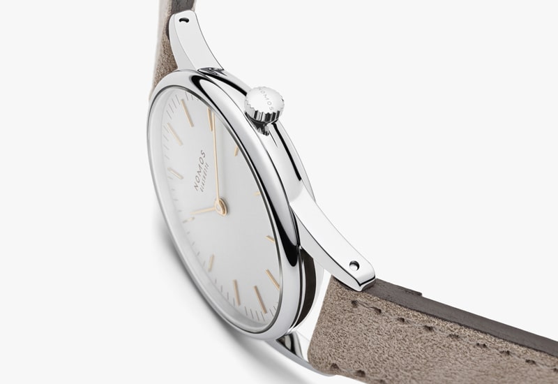 Juwelier-Wagner-Madler-Uhren-Nomos-Glashuette-Orion-Kollektion-Ansicht-1 Eine elegante Uhr aus der Nomos Orion Kollektion mit silbernem Gehäuse und Krone, minimalistischem weißen Zifferblatt und goldenen Stundenmarkierungen. Sie hat ein hellbraunes Lederarmband und ist so abgewinkelt, dass sich ihr schlankes Profil vor einem weißen Hintergrund abhebt.