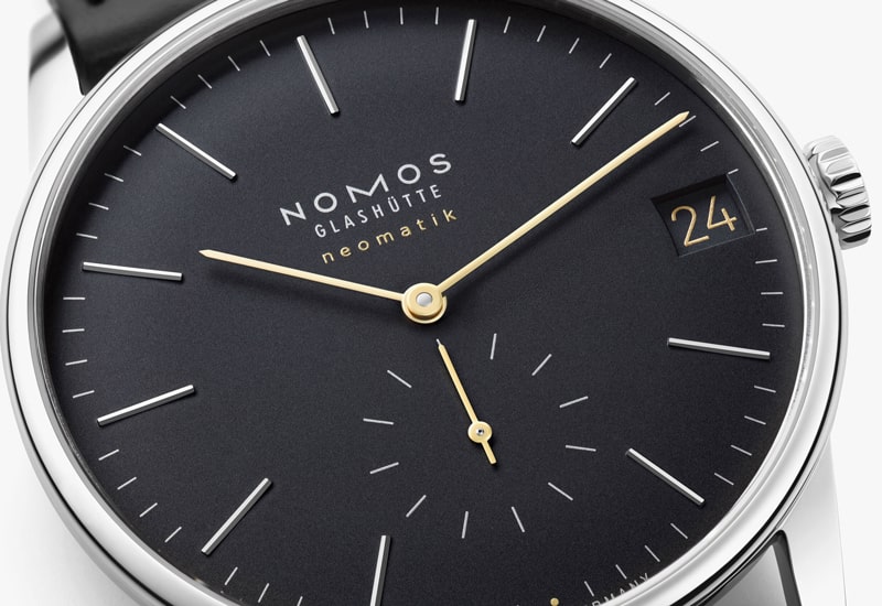 Juwelier-Wagner-Madler-Uhren-Nomos-Glashuette-Orion-Kollektion-Ansicht-2 Nahaufnahme einer Uhr von Nomos Glashütte aus der Orion Kollektion mit einem eleganten schwarzen Zifferblatt. Sie verfügt über minimalistische goldene Zeiger, schmale Stundenmarkierungen, ein zweites Hilfszifferblatt und ein Datumsfenster mit der Anzeige „24“, alles angetrieben vom automatischen Uhrwerk neomatik.