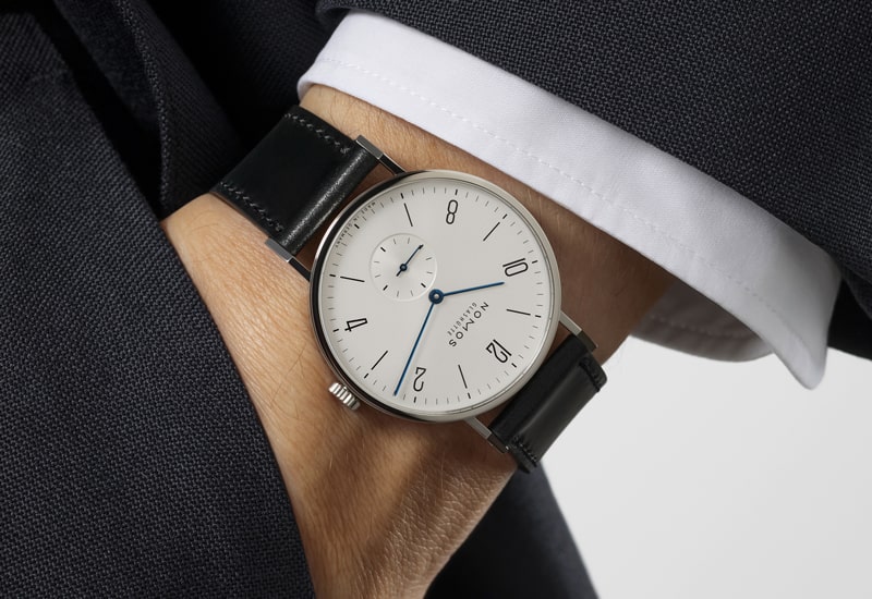 Nahaufnahme einer Person, die eine elegante Armbanduhr mit schwarzem Lederarmband aus der Nomos Tangente Kollektion mit weißem Zifferblatt, schwarzen Ziffern und blauen Zeigern trägt, elegant unter die Manschette eines dunklen Anzugs und eines weißen Hemdes gesteckt.
