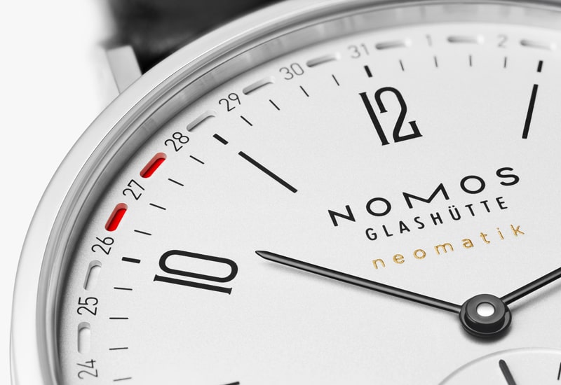 Nahaufnahme eines Zifferblatts der Nomos Glashütte neomatik aus der angesehenen Tangente Kollektion. Das Design zeichnet sich durch ein minimalistisches weißes Zifferblatt mit schwarzen Stundenmarkierungen, goldenem Markentext und einer Kalenderanzeige mit roter Markierung neben der Zahl 27 aus.