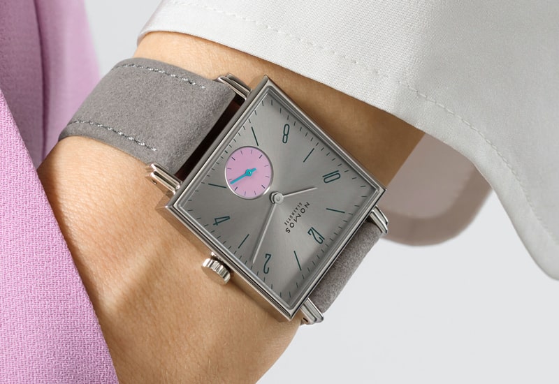 Juwelier-Wagner-Madler-Uhren-Nomos-Glashuette-Tetra-Kollektion-Ansicht-1 Eine quadratische Armbanduhr aus der Nomos Tetra Kollektion mit grauem Zifferblatt und blauen Ziffern ziert das Handgelenk einer Person. Sie verfügt über ein kleines rosa-blaues Hilfszifferblatt und ein graues Wildlederarmband. Die Person ergänzt die Uhr wunderbar mit ihrem weiß-rosa Outfit.