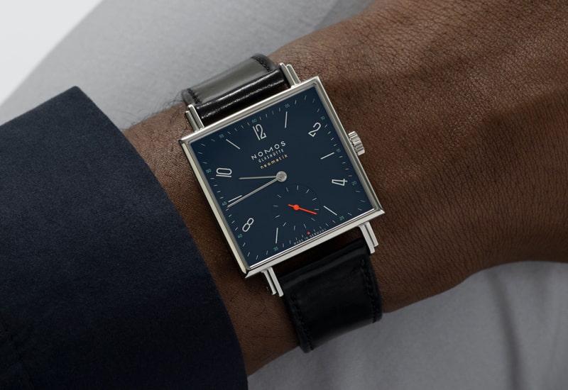 Juwelier-Wagner-Madler-Uhren-Nomos-Glashuette-Tetra-Kollektion-Ansicht-2 Ein Handgelenk, das eine quadratische, schwarze Uhr mit Lederarmband aus der Nomos Tetra Kollektion mit dunkelblauem Zifferblatt und minimalistischem Design trägt. Die Uhr hat silberne Stundenmarkierungen, schlanke Zeiger und einen roten Sekundenzeiger. Der Träger trägt einen dunklen Hemdsärmel.