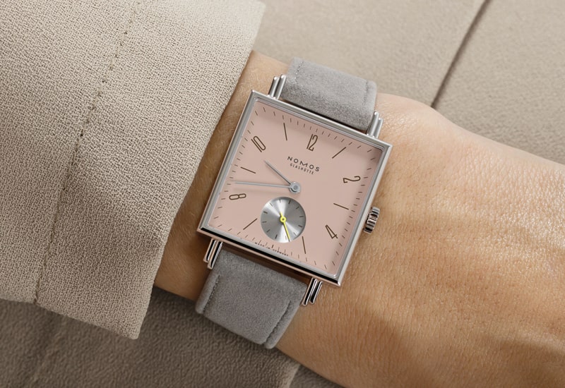 Juwelier-Wagner-Madler-Uhren-Nomos-Glashuette-Tetra-Kollektion-Ansicht-3 Eine quadratische Uhr aus der Nomos Tetra Kollektion mit hellrosa Zifferblatt und schlanken silbernen Zeigern schmückt das Handgelenk. Sie verfügt über ein graues Lederarmband und ein kleines silbernes Hilfszifferblatt auf beigem Stoffhintergrund.