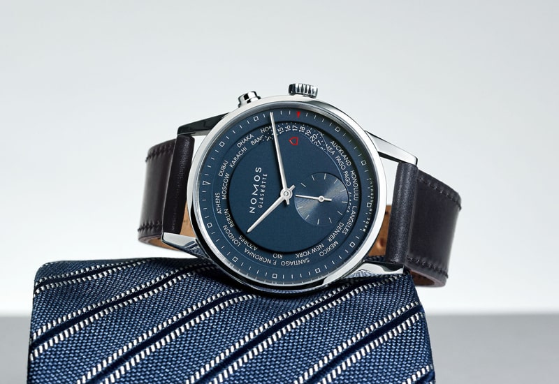 Juwelier-Wagner-Madler-Uhren-Nomos-Glashuette-Zuerich-Kollektion-Ansicht-1 Eine elegante Armbanduhr aus der Nomos Zürich Kollektion mit blauem Zifferblatt, das Zeit und Datum anzeigt, ruht anmutig auf einer blau-weiß gestreiften Krawatte. Das elegante Design wird durch das silberne Gehäuse und das dunkle Lederarmband hervorgehoben.