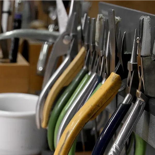 Juwelier-Wagner-Madler-Mainz-Schmuck-Atelier-Goldschmiede-3 Eine Nahaufnahme mehrerer Zangen mit verschiedenfarbigen Griffen, die an einem Gestell hängen. Im Hintergrund sind weitere Werkzeuge und ein weißer Behälter zu sehen, der leicht unscharf ist. Das Bild lässt auf eine Goldschmiedewerkstatt oder einen Werkzeuglagerbereich schließen.