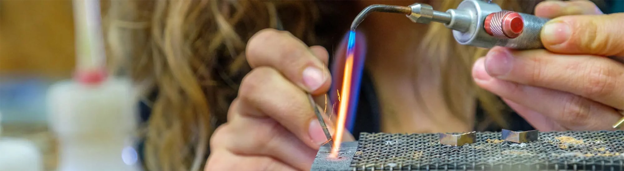 Nahaufnahme von Händen, die mit einer kleinen Fackel mit blauer und orangefarbener Flamme an einem Schmuckstück aus Metall arbeiten. Die Person hält ein Metallstück mit einer Pinzette über einer Werkbank mit perforierter Oberfläche. Auf der Werkbank sind Ringe und Werkzeuge zu sehen.