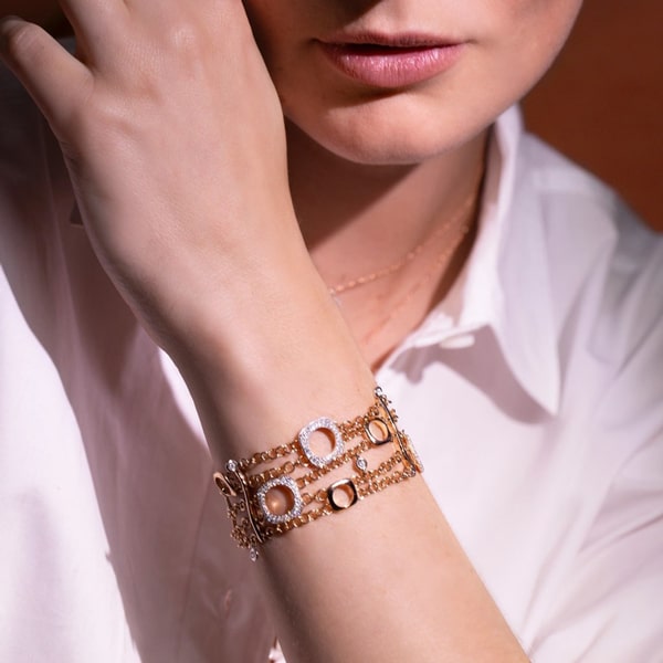 BIGLI © 2024 Eine Person in einem weißen Hemd trägt elegante Bigli Armschmuck-Armbänder mit kreisförmigen Mustern an ihrem Handgelenk. Diese Armbänder kombinieren Gold- und Silberelemente und reflektieren das Licht auf subtile Weise. Die Lippen und das Kinn der Person sind sichtbar, was den anspruchsvollen Reiz noch verstärkt.