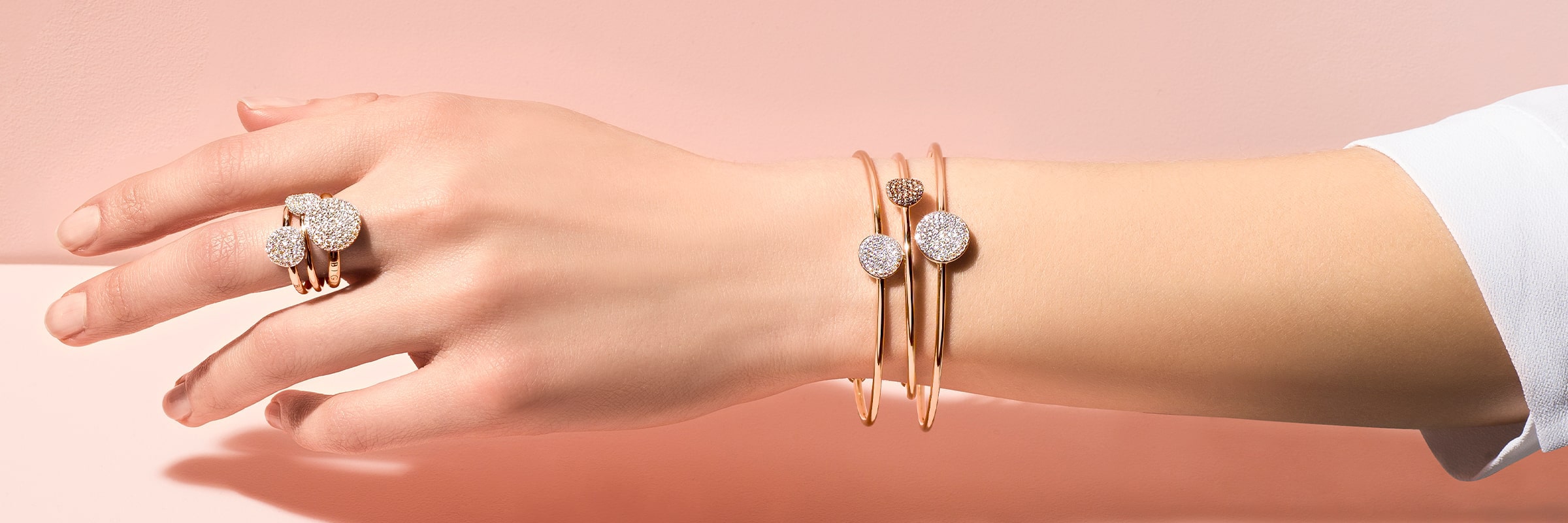 Eine Hand trägt zwei Armreifen aus Roségold mit diamantbesetzten runden Enden und einen dazu passenden Ring mit drei diamantbesetzten Kreisen vor einem hellrosa Hintergrund.