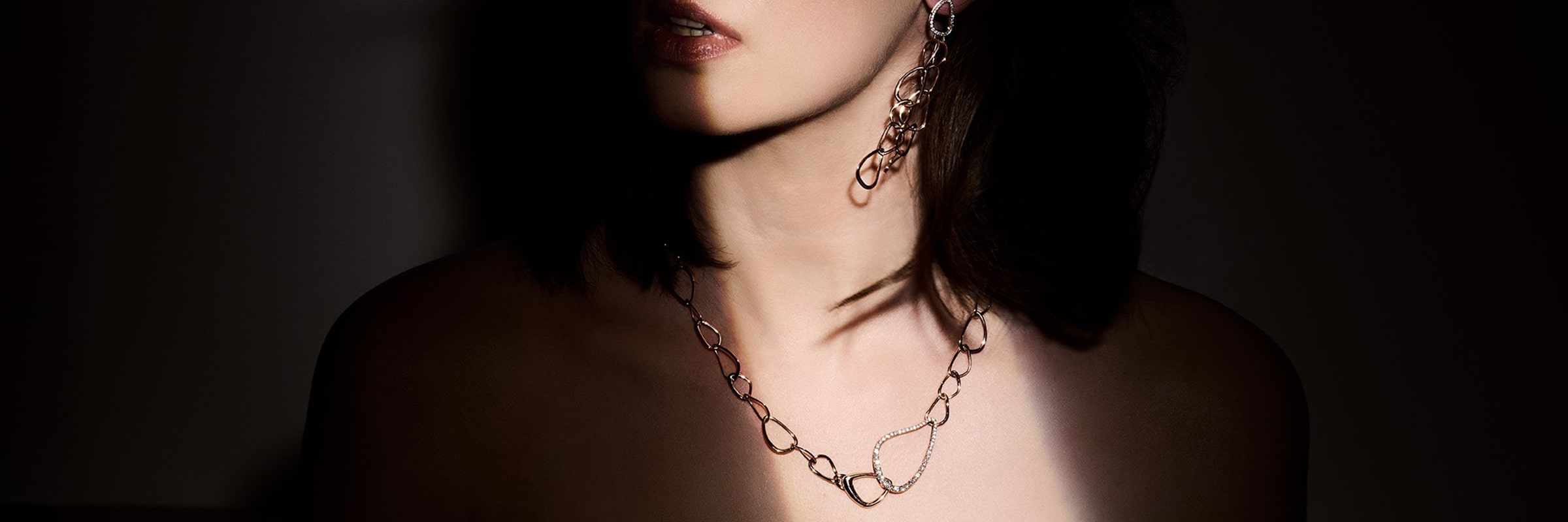 Eine Person trägt eine silberne Halskette und dazu passende Ohrringe. Das Bild ist kunstvoll beleuchtet, um den Schmuck hervorzuheben und Schatten auf das Gesicht und die Schultern der Person vor einem dunklen Hintergrund zu werfen.